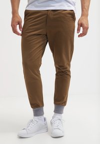 Pantaloni jogger marroni con taglio affusolato, realizzati in tessuto morbido con polsini elastici, abbinati a sneakers bianche e calzini alla caviglia grigi.