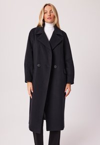 Długi czarny wełniany płaszcz z klapami, zapinany na podwójne guziki, z bocznymi kieszeniami; charakteryzuje się luźnym krojem i delikatną fakturą.