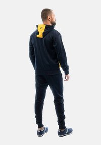 Tuta di colore navy con dettagli gialli sulla cappuccio, realizzata in tessuto morbido. Presenta una vita elastica e polsini elasticizzati, abbinata a scarpe da ginnastica blu.