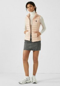 Light pink puffer vest with a zip closure and pockets, worn over a white ribbed turtleneck, paired with a denim skirt and silver sneakers.
