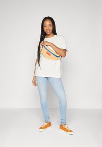Svijetlosiva oversized majica, plave izgrebane skinny traperice, narančasti tenesici i bež bum bag s plavim patentnim zatvaračem.