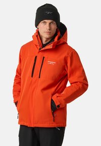Orange insulated jacket with a detachable hood, black accents, and zippered pockets. Model wears a black knit beanie. Textured fabric.