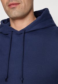 Hanorac bleu închis cu glugă, realizat dintr-un material texturat, cu șnururi și un design simplu. Prim-plan care arată zona gâtului și detaliile glugii.