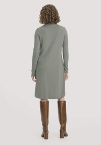 Knielanges, langärmeliges Kleid aus mattem grünen Stoff mit glatter Textur. Kombiniert mit braunen kniehohen Stiefeln und einem schlichten Ausschnitt.