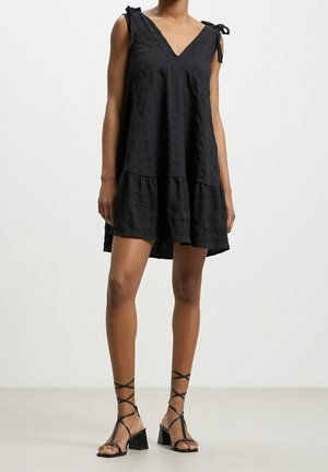 Kobieta w czarnej, bezrękawnikowej sukience o fakturze, z dekoltem w serek i wiązaniami na ramionach, połączonej z czarnymi sandałami na obcasie typu blok z wiązaniem na sznurowadła.