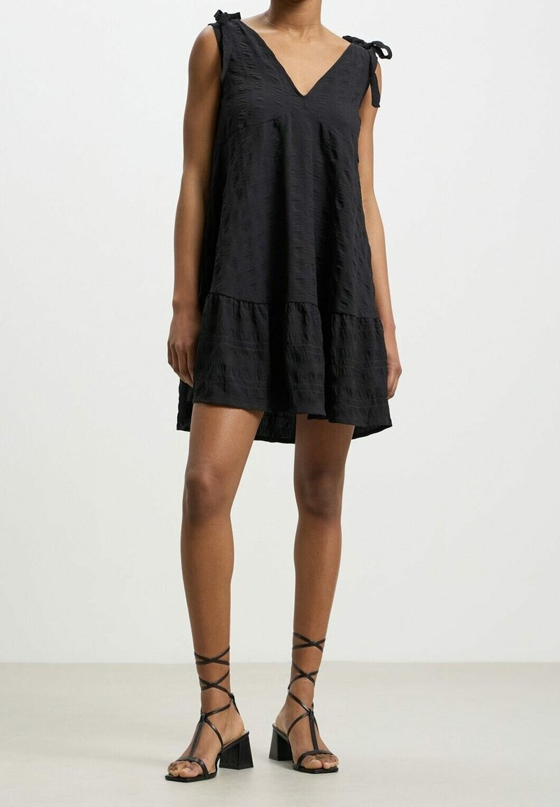 Femme portant une robe noire texturée sans manches avec un décolleté en V et des liens aux épaules, associée à des sandales à talons blocs noires à lacets.