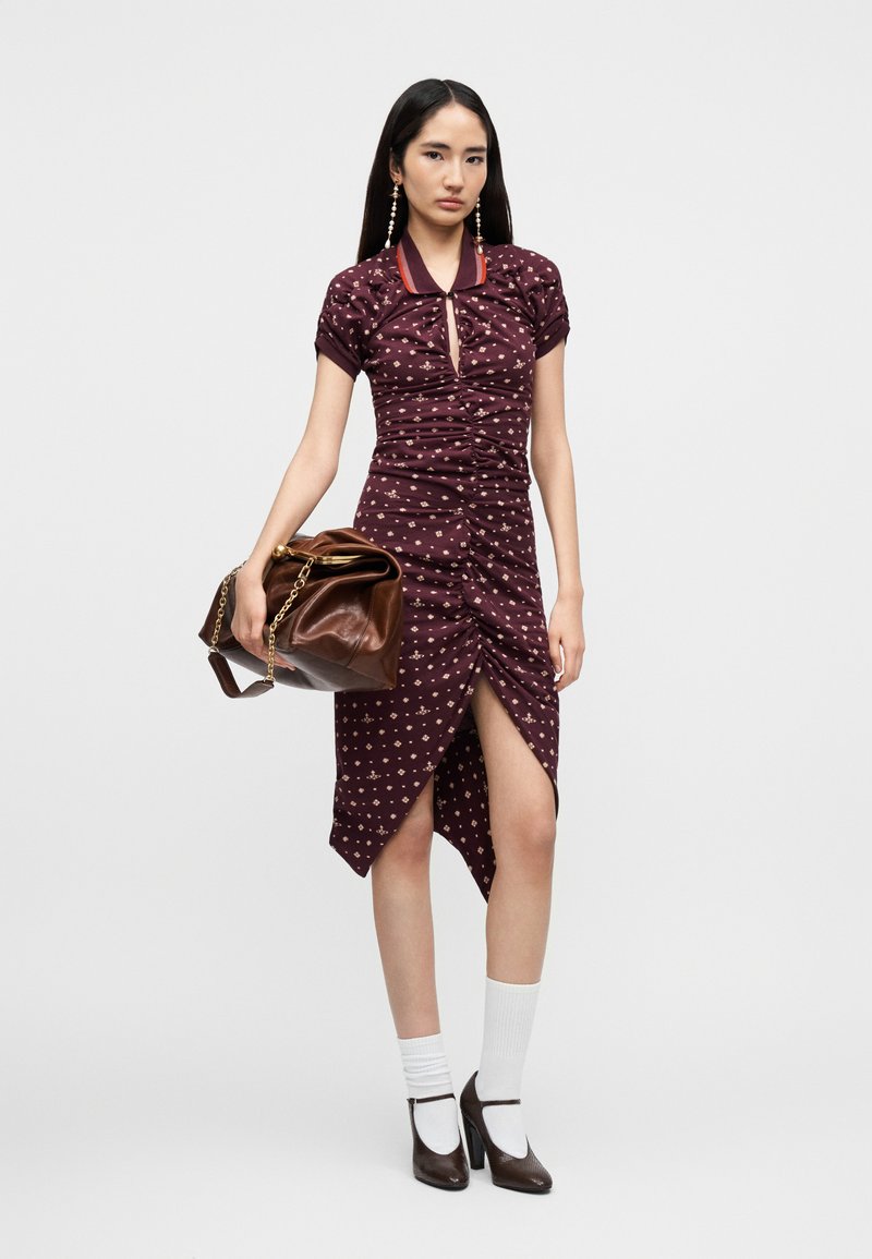 Bordeauxrotes Kleid mit kurzen Ärmeln, Rüschendetails, kleinem Blumenmuster und hohem Schlitz. Kombiniert mit einer braunen Handtasche und Blockabsatzschuhen.