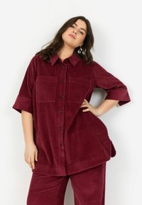 Camicia in velluto a coste bordeaux con chiusura a bottoni, due tasche sul petto, maniche corte e una vestibilità leggermente over. Tessuto testurizzato.