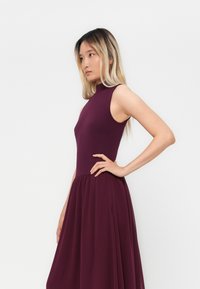 Young woman with long blonde hair wears sleeveless high-neck purple dress, standing sideways with hand on hip against plain white background.