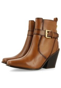 Botas de tobillo de cuero marrón con puntera puntiaguda y tacón cuadrado. Presenta una correa con una hebilla dorada y detalles de costura en los laterales.
