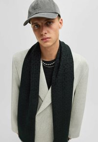 Sciarpa a motivo verde scuro con una texture morbida, indossata con un blazer grigio chiaro. Accentuata da una catena d'argento a strati e un cappellino grigio.