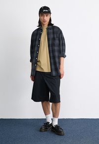 Model wearing a black and grey plaid button-up shirt over a beige t-shirt, black shorts, white socks, and black shoes with thick soles.