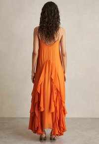 Dlouhé, oranžové, splývavé šaty s vrstvenými volány na zadní straně, tenkými ramínky a volnou, lehkou tkaninou. Měkká textura a elegantní silueta.
