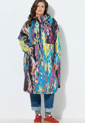 Femme en imperméable à capuche avec un motif abstrait coloré, jean bleu retroussé, chaussettes rayées et baskets rouges, debout devant un fond blanc.