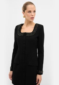 Robe noire avec un décolleté carré, un col et des poignets ornés. Présente des boutons sur le devant et deux poches latérales. Texture de tissu lisse.