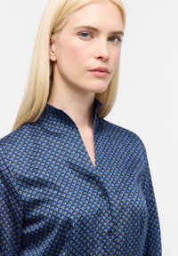 Blusa blu a motivi con colletto alto, caratterizzata da un design geometrico in tonalità più chiare. Tessuto morbido con chiusura a bottoni sul davanti.
