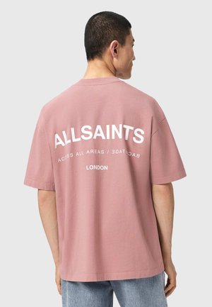 Mężczyzna w niemalowym, różowym oversize'owym t-shircie z nadrukiem "ALLSAINTS" i "LONDON" w białym kolorze na plecach, w połączeniu z jasnoniebieskimi dżinsami.