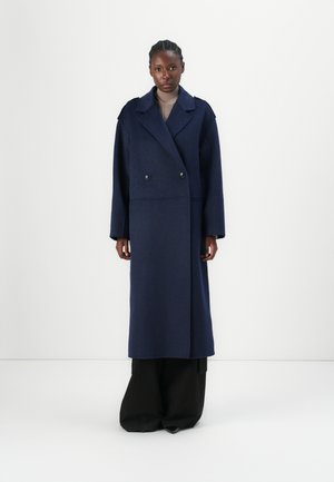 Double-breasted long coat in dark navy wool blend, featuring large notched lapels, dropped shoulders, and a relaxed fit, paired with wide black trousers.