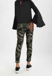 Kamouflagebyxor i grön, svart och beige med en figurnära design. Tillsammans med en svart topp med trumpetärmar och ett nyckelhål bak.