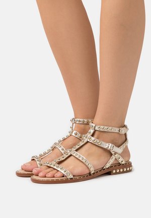 Sandalen met bandjes en metalen studs, met een tan leren zool en verstelbare enkelbandjes. De kleur is lichtbeige met een gladde textuur.