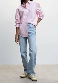 Femme vêtue d'une chemise à rayures rose à boutons avec les manches retroussées, un jean droit bleu clair et des baskets beige, debout devant un fond uni.