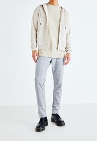 Felpa beige con zip sopra una maglietta color sabbia chiaro, abbinata a pantaloni grigi a quadri. Calzature: scarpe nere con suola rialzata e cuciture gialle.