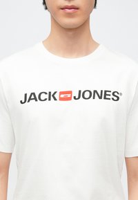 Camiseta blanca de algodón con un logo negro y un acento rojo, mangas cortas y diseño clásico de cuello redondo.