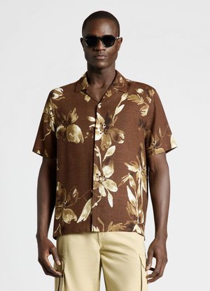 Mann mit dunkler Sonnenbrille, braunem kurzärmeligem Hemd mit großem beigem Blumenmuster und hellbeiger Hose vor einfarbigem Hintergrund.