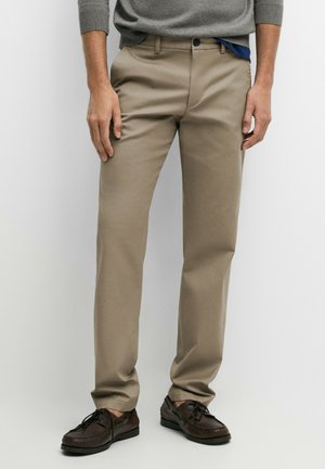 Homme portant un pantalon beige droit, des chaussures à lacets marron foncé et un pull gris, debout devant un fond clair uni.