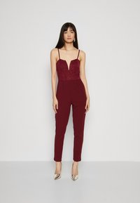 Burgunderfarget jumpsuit med blondebodice, dyp V-hals, tynne stropper og avsmalnende ben. Kombinert med metalliske høye hæler.