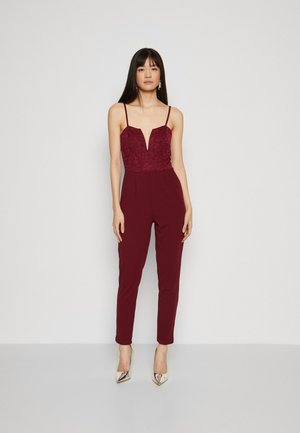 Burgunderfarget jumpsuit med blondebodice, dyp V-hals, tynne stropper og avsmalnende ben. Kombinert med metalliske høye hæler.