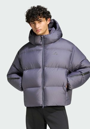 Giovane uomo con capelli ricci, indossa una giacca grigia imbottita con cappuccio e pantaloni neri, con le mani nelle tasche.