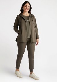 Kvinna som bär en olivgrön zip-up hoodie, matchande byxor och en t-shirt med pärlbeslag, tillsammans med vita sneakers, står och ler.
