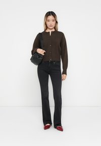 Bruine gebreide cardigan met knopen, gecombineerd met zwarte flared jeans en rode puntige schoenen. Model houdt een kleine zwarte tas vast.