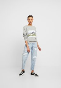 Sweatshirt cinza com uma estampa de montanha, punhos e bainha canelados. Combinado com jeans claros desgastados e ténis de atacadores em preto e branco.