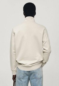 Persona con un sudadera color crema y pantalones vaqueros de un azul claro, de pie mirando hacia un lado con una mano en el bolsillo.