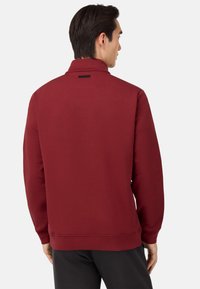 Bordo pulover s visokim ovratnikom, rebrastimi manšetami in robom. Ima teksturirano blago in majhno nalepko blagovne znamke na hrbtu.