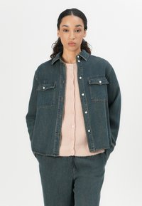 Dunkelblaue Jeansjacke mit zwei Brusttaschen und silbernen Knöpfen, übereinander getragen mit einem weichen, rosa gerippten Pullover.