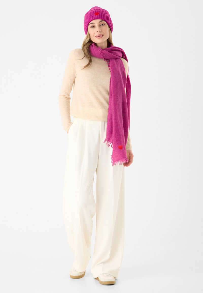 Geribbeld roze beanie met harten-detail, beige trui, felroze sjaal met franjes, brede witte broek en crèmewitte sneakers.