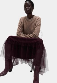 Kobieta siedząca na białym krześle, ubrana w brązowy sweter z ozdobnym dekoltem, ciemnofioletową tiulową spódnicę i dopasowane wysokie buty.