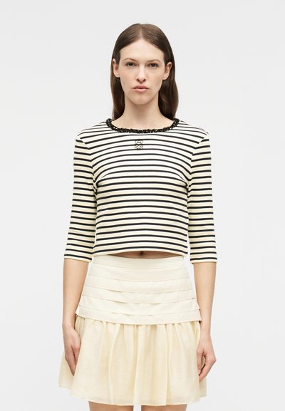 Γυναίκα με ίσια καστανά μαλλιά φοράει μαύρο-λευκό ριγέ κοντό τοπ με τρία τέταρτα μανίκια και κρεμ φούστα με βολάν.