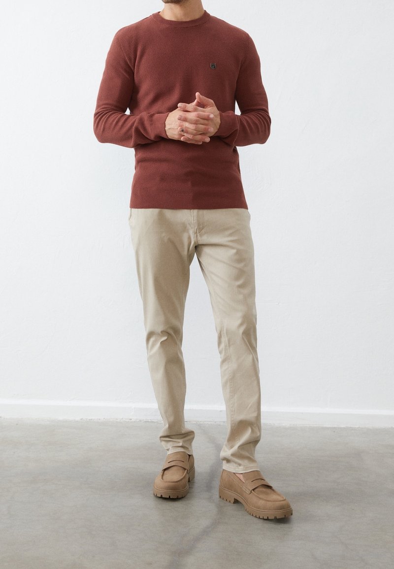 Pull en tricot de couleur rouille avec un col rond, associé à un chino beige et des chaussures à enfiler marron avec des semelles texturées.