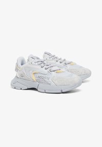 Sneakers Lacoste con una parte superiore in rete e suede grigio chiaro, dettagli gialli e una suola in gomma spessa. Presentano un design con lacci e un logo distintivo.