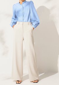 Blusa azzurro chiaro con colletto a balze e chiusura a bottoni, abbinata a pantaloni ampi color crema e sandali neri con lacci.