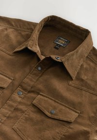 Chemise en velours côtelé marron avec poche poitrine à rabat boutonné, boutons-pression et col pointu, posée à plat sur une surface claire.