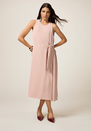 Lichtroze, mouwloos jurkje met een zijstrikdetail, vloeiende stof en een rechte silhouette. Gekoeld met bordeauxkleurige puntige flats.