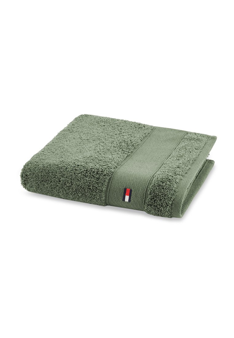 Serviette en coton vert avec une surface douce et texturée. Présente une bande lisse et un petit logo de marque en rouge, blanc et bleu sur le bord.