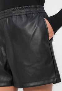 Czarne szorty z imitacji skóry ze ściągaczem w pasie, wyposażone w dwie boczne kieszenie i gładką fakturę, częściowo widoczne z boku.