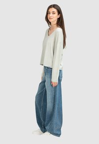 Hellgrauer, langärmeliger V-Ausschnitt-Pullover kombiniert mit weit geschnittenen blauen Jeans und weißen Sneakers, die einen entspannten Schnitt und einen lässigen Stil präsentieren.