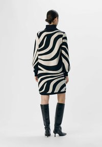 Robe pull rayée noir et crème avec un col montant, des épaules basses et une silhouette ajustée, associée à des bottes noires montantes.